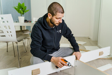 One Man with beard alone Putting Together Self Assembly Furniture at Home holding electric screwdriver and plywood parts with screws - half length front view DIY concept real people copy space