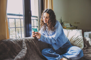 young woman looking at the mobile on the sofa near the window