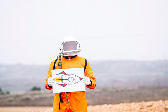Man dressed as an astronaut holds a poster with a drawing of a rocket