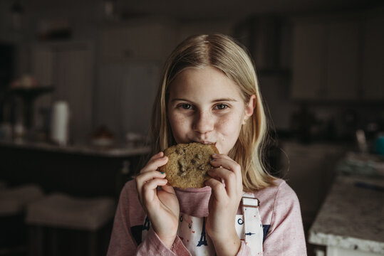 Tween Girl Eating A Large Chocolate Chip Cookie In Kitchen With Apron