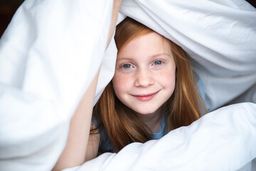 Young red haired girl peeking out from under a white comforter.