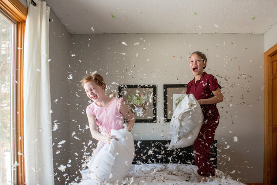 Two Happy Young Girls Having A Feather Pillow Fight On The Bed.