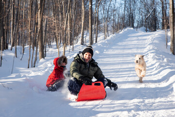 Father and son sledding down snowy hill with their dog on winter day.
