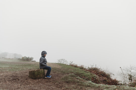 Boy sitting on concrete post in winter coat against foggy overcast sky