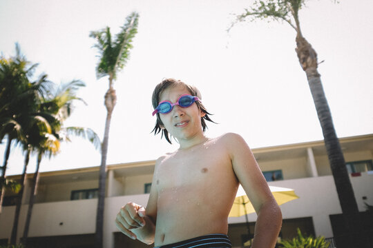 Boy in Goggles with Wet Hair stands Pool Side with Palm Trees