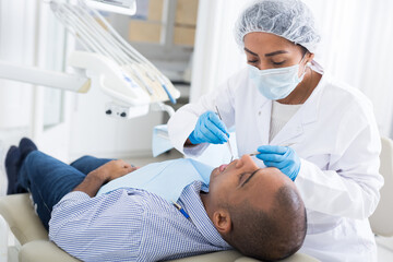 Confident hispanic female dentist treating teeth of male patient in modern dental office