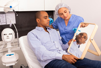 Confident cosmetologist examining Hispanic male client face before procedure in esthetic clinic. Man looking in mirror