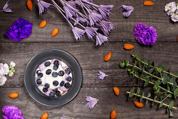 Obraz na płótnie Canvas Blueberry Graham Cheesecake topped with fresh berries on a dark wood background with almonds, purple flowers and twigs. Flat lay, top view.