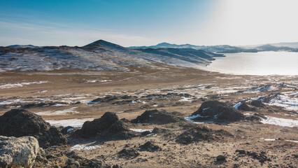 The frozen lake is surrounded by hills. The snow is lying on the bare ground. Picturesque boulders in the foreground. Blue sky. Baikal.