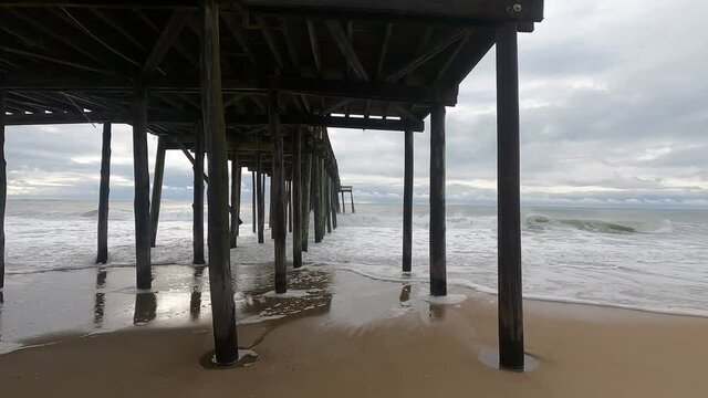 Sea foam creeps up the beach even as waves of salty ocean water retreat back into the sea, seen from underneath a wooden fishing pier along the coast of a tourist destination town