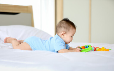 Adorable and innocent little Asian baby enjoys playing fun with colorful children toy while lying on stomach on bed.
