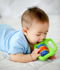 Adorable and innocent little Asian baby enjoys playing fun with colorful children toy while lying on stomach on bed.