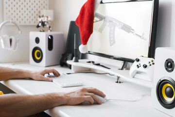 Male hands on the computer table hold a gaming mouse and keyboard. A red Santa hat hangs on the monitor. 