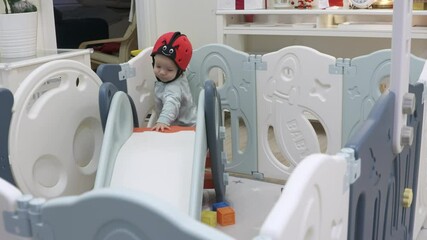 Cute toddler in baby safety helmet enjoy playing time in infants playpen with slide, 8 month old baby boy playing in safe activity center. High quality 4k footage