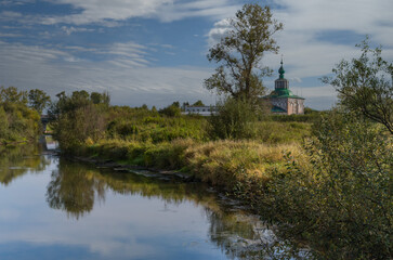 Fototapeta na wymiar Summer landscape in the city limits of Solikamsk (Russia). The raised bank of the river, overgrown with grass, trees with kutarniks, which are reflected in the water. In the distance the old church
