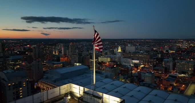 American flag waves in wind at night in urban America USA city. Patriotic theme at sunset. Aerial view.
