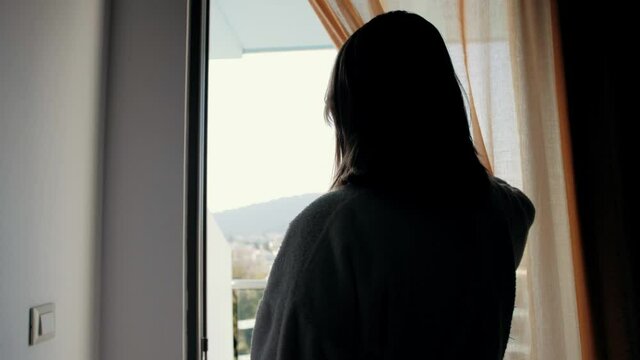 Woman in hotel room opens curtains and walks balcony, beautiful city view.