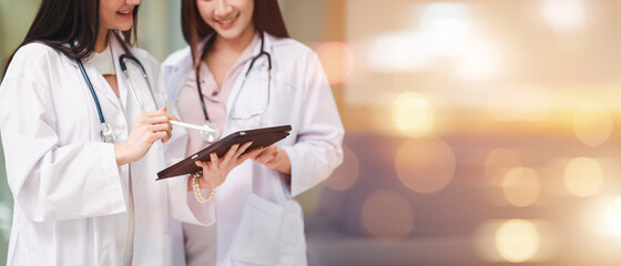 Doctor in a uniform, a stethoscope and a female assistant stand together in a hospital corridor. Hold the data file and tablet showing the patient's symptoms. Asian and European women.