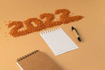 The number 2022 next to a notebook and a planning pen. The figure is made of bright orange shiny beads. Signs and symbols of the new year for copy space. Festive mockup.