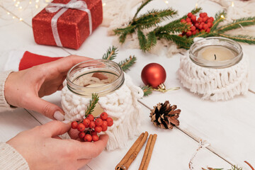 A woman decorates a glass candlestick in the macrame style weaving,fir branches and rowan berries.Handmade decor in eco-style,hands close-up.Christmas, New Year and eco-friendly concept.