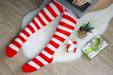 Girl in Christmas knee socks sitting on a fur rug with laptop near the gift boxes. Ordering gifts, work and leisure at home during New Year holidays