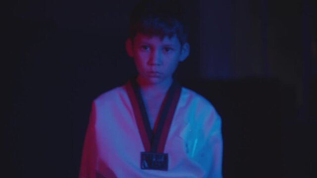 A little boy doing taekwondo in the dark with neon lighting - walks to the camera and showing his hands in gesture