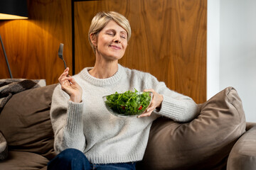 Middle-aged woman is sitting and resting at home on the couch, eating a green salad. A woman eating...