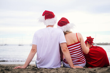 Obraz premium back view Happy family in Santa Claus hats celebrating Christmas on beach. people traveling to warm countries on winter tour. New Year and Christmas tourists