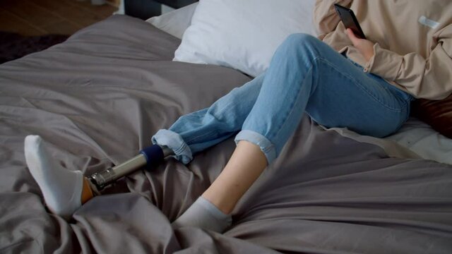 Cute dreamy young female with congenital disability and prosthetic leg sitting on bed, enjoying music in wireless headphones using mobile phone app while relaxing in domestic room.