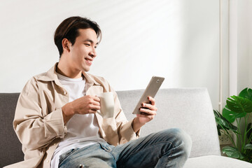 Happy Asian student sits and relaxes with a cup of coffee or tea using modern tablet browsing unlimited wireless internet, young man freelance working uses wifi at a home apartment in the morning
