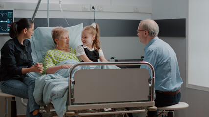 Kid hugging sick grandma at family visit in hospital ward. Mother, little girl and old man talking...