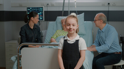 Fototapeta premium Portrait of kid in hospital ward visiting aged patient in bed to give support. Child smiling at family visit for senior woman with disease, giving assistance to grandma with mother and old man.