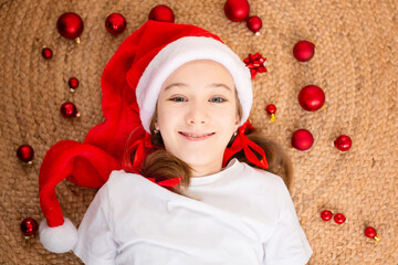 Cute girl in Santa Claus hat are lying on the floor among red Christmas balls at home. top view. Christmas. A time of miracles and fulfillment of desires. Merry Christmas.