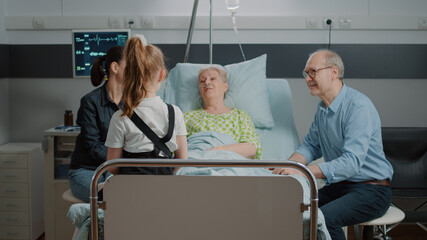 Retired patient talking to niece at family visit in hospital ward. Child, mother and old man visiting person with illness in bed at intensive care. Adult having conversation with visitors.