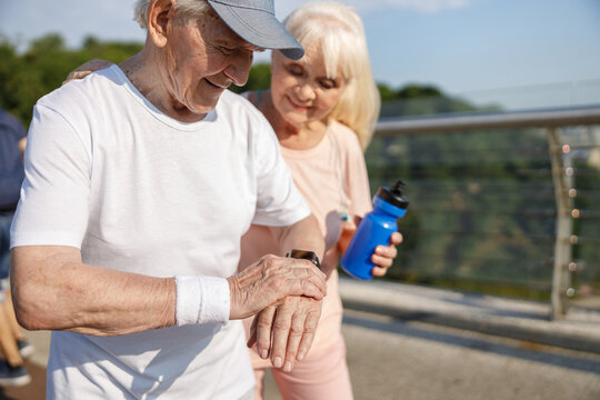 Senior man with cap monitors heart rate with smartwatch at training with wife on footbridge