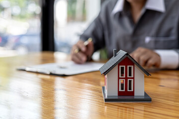 Replica of a small orange house sits on a table, a housing project salesperson is drafting a sales contract for a customer who reserves a house in the project he maintains. Real estate trading concept