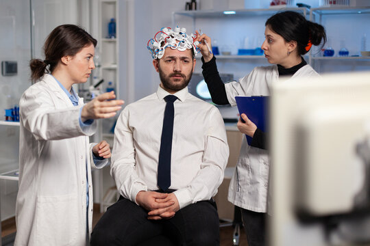 Medical Team Of Scientists Adjusting Brain Eeg Analyzing Nervous System Evolution Of Man Patient During Medicine Experiment In Neuroscience Laboratory. Physician Doctor Monitoring Neurology Activity