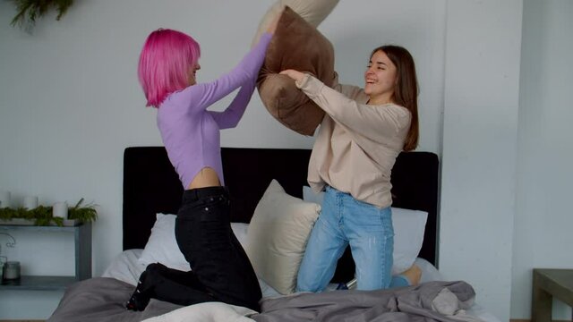 Joyful Stylish Attractive Pink Haired Young Woman And Excited Smiling Cute Female Roommate With Birth Hands Anomaly And Artificial Limb Enjoying Leisure, Having Pillow Fight On Bed In College Dorm.