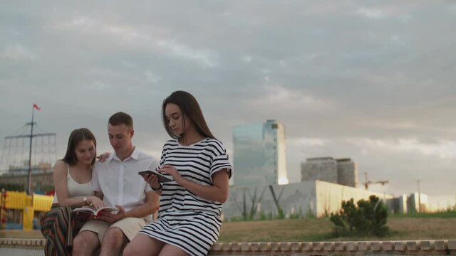 Two girls and a guy sitting in the park during the day reading an e-book