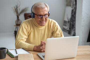 Old man enjoying pleasant chat while working on laptop with headphones
