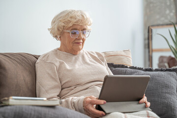 Senior woman wearing glasses sitting at the couch at home and using tablet