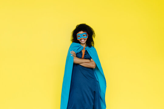 Smiling woman wearing eye mask and cape standing against yellow background