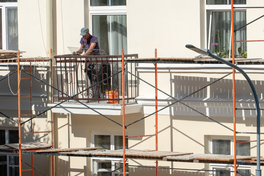 Repair of the balcony and facade of the building. The builder stands on the balcony and holds a sheet of glass and tools in his hands. Scaffolding is located along the walls of the building.