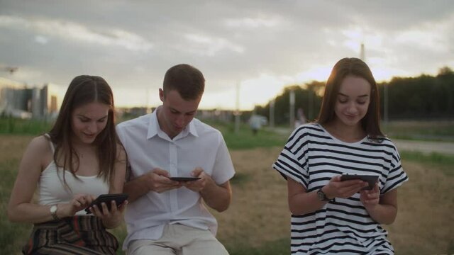 Two girls and a guy sitting in the park during the day reading an e-book
