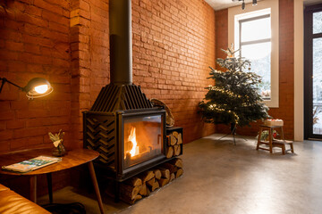 Cozy living room interior with Christmas tree and burning fireplace. Concept of home comfort in winter. Windows overlooking on snowy garden, room with brick wall and concrete floor