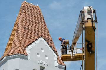 Repair and reconstruction of the old roof made of red tiles of a century-old building. The worker is in the basket of a crane and works at a height in dangerous conditions. Roof repair on a sunny day