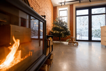 Cozy living room interior with Christmas tree and burning fireplace. Concept of home comfort in winter. Windows overlooking on snowy garden, room with brick wall and concrete floor