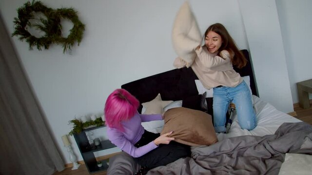 Portrait of carefree happy attractive young woman with congenital disability and prosthetic leg and cheerful pretty pink haired female friend having fun, pillow fighting on bed in college dorm room.