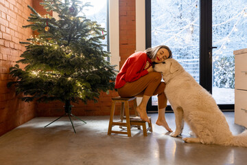 Woman hugs with a dog, enjoying winter holidays at home. Living room interior with Christmas tree and windows ovelooking on a snowy garden