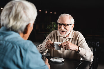joyful senior man in eyeglasses holding coffee cup near blurred friend in bar
